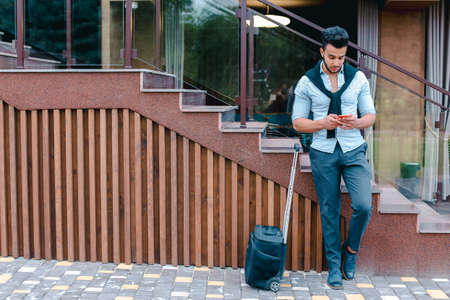 Young man businessman Arabian beautiful appearance is black travel bag on small wheels and is waiting for taxi, on journey or business trip, using phone and check their seats in airplane online, reserve hotel, one leg held up other, and based on stairs near restaurant on open air. Handsome Muscular Guy Dressed in Black Classic Trousers, White Shirt on Top of Which Knotted Black Sweater. Concept of Business Meeting, Airport, Flight Has Been Delayed, Decides to Explore New Places.の写真素材