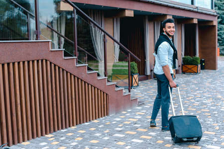 Young Guy Arab Businessman Student Came go to Restaurant With Suitcase in Hand, Smiling at Camera, Stops and Looks Away.の写真素材