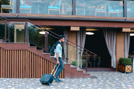 Young Guy Arab Businessman Student Came go to Restaurant With Suitcase in Hand, Smiling at Camera, Stops and Looks Away.の写真素材