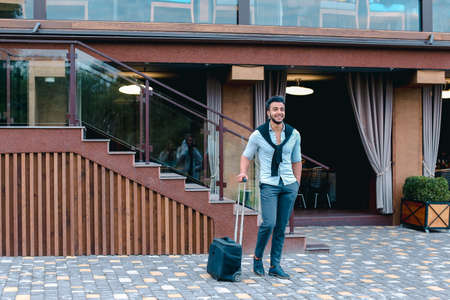 Young Guy Arab Businessman Student Came go to Restaurant With Suitcase in Hand, Smiling at Camera, Stops and Looks Away.の写真素材