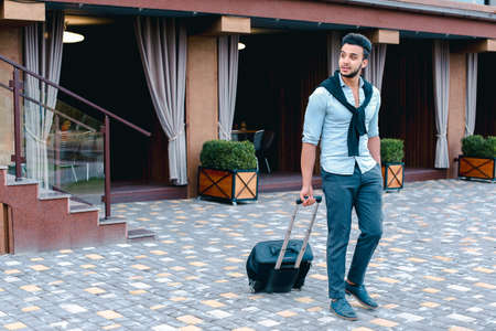 Young Guy Arab Businessman Student Came go to Restaurant With Suitcase in Hand, Smiling at Camera, Stops and Looks Away.の写真素材