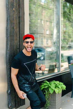 Handsome young man arab stands near wall smiles, rejoices and holds in hand mobile phone on background of windows in modern restaurant.の写真素材