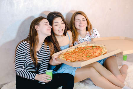 Cute girls cool spend time and enjoy pizza, sit on floor in bright bedroom in evening.の写真素材