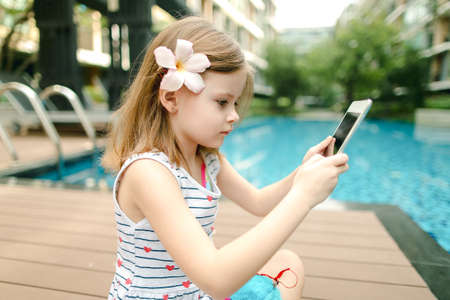 little girl uses tablet sitting close to swimming pool wearing fの写真素材