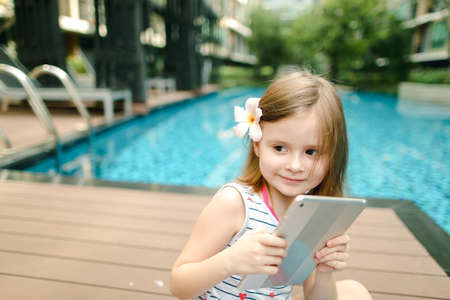 little kid taking selfie on tablet sitting close to swimming pooの写真素材