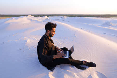 Handsome young Arab man writes on Internet using laptop and sits on sand amid sandy desert at sunset.の写真素材