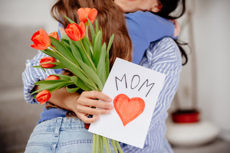 Grateful mom hugging daughter girl, holding flowers bouquet, receiving hand drawn greeting card with loving heart from girl, smiling at camera. Mothers day, 8 march, concept. Head shot portrait. Close-up of hands holding a bouquet of tulips and a handmade cardの写真素材