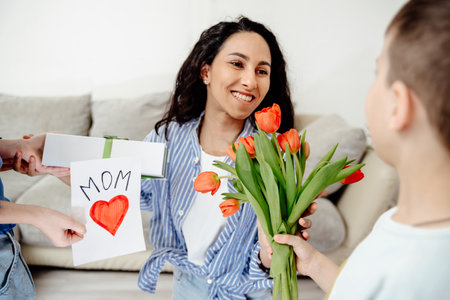 Arab Mom receives gifts from her children. The son holds out a bouquet of tulips and the daughter a handmade card and a gift. Everyone is in a bright room in a cozy homely atmosphere.の写真素材