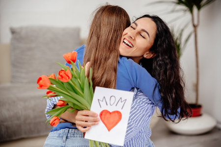 Child daughter congratulates mom and gives her flowers tulips and postcard. Mum and girl smiling and hugging. Family holiday and togetherness. Mom hugs her daughter in her arms, holds her gifts and closed her eyes in pleasureの写真素材