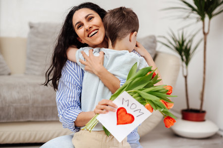 Little boy greeting his mom on Mother's Day at home. The son gave his mother a bouquet of flowers and a handmade card.Mom hugs her child and smiles broadly.の写真素材