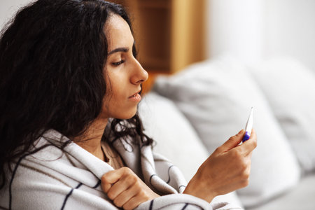 A young beautiful Arab woman holds a thermometer with one hand and holds a plaid with the other while sitting on a gray sofa. A woman looks puzzled at a thermometer in a homely atmosphere.の写真素材