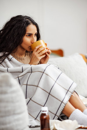 Beautiful young Arab girl drinks a cup of hot drink. Woman is wrapped in a blanket and sits on a gray sofa near her there are a lot of medicines. Recovery at home is always comfortable and safe.の写真素材