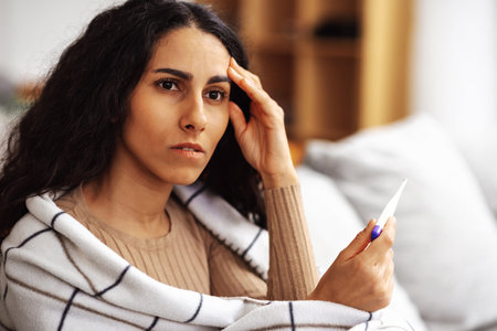 Young beautiful Arab woman falls ill and looking ahead thoughtfully. Woman holds a thermometer with one hand and she holds her head with her other hand with the other while sitting at home.の写真素材