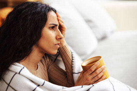 Young beautiful woman of Arab is sitting on a gray sofa wrapped in a blanket in one hand holding a cup with the other hand holding her head. Woman coping with stress and depression problems.の写真素材