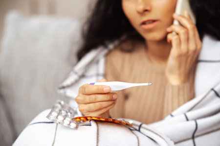 Woman sits in her room on a gray sofa wrapped in a blanket. She is ill and the call to the doctor is a consultation about her treatment. Hand has a phone. Other hand an electronic thermometer.の写真素材