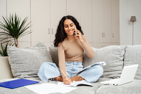 Young beautiful female student of Arab appearance sits on the sofa at home and studies. She holds a phone in one hand and takes notes in the other. Carries on a pleasant conversation about the subject of the task.の写真素材