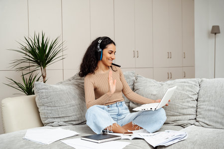 A young beautiful girl of Arab appearance leads a video call. Greeting waving hand and holding a laptop. The girl is sitting on the couch in a homely atmosphere around her a lot of papers.の写真素材