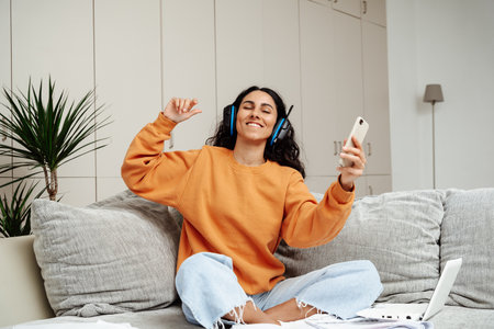 A young beautiful girl of Arab appearance sits on a sofa in a homely atmosphere. The girl dances and listens to music in headphones enjoys a break in the working moment.の写真素材
