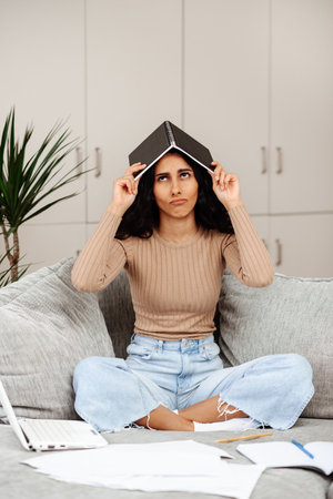 Indoor shot of tired woman wearing casual clothing sitting on sofa and working on notebook, felling fatigue. She covered herself with a notebook and hides like a house from a large amount of paperworkの写真素材