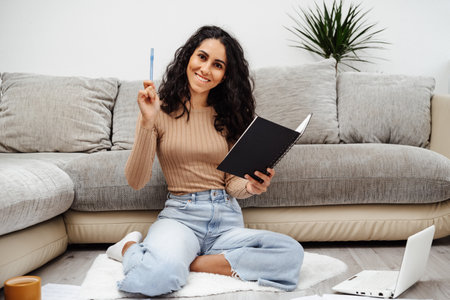 Young beautiful Arab woman holds a notebook in her hand and makes a wish list. She took a pen and is ready to paint everything point by point. The girl sits on the floor and smiles brightly at home.の写真素材