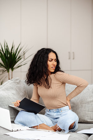 Portrait of tired exhausted Arab woman sitting on sofa and working, spending long hours in front of laptop and notepad, feeling back pain.の写真素材
