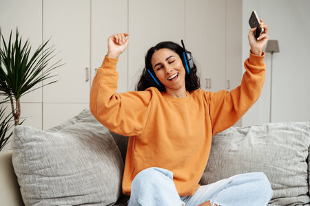 Carefree brunette listening to music and singing, using headphones, sitting on the sofa in the living room, copy space. Happy young lady with closed eyes enjoying music, home interior.の写真素材