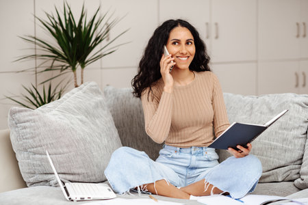 Smiling arabic millennial woman talking on phone holding note book sitting on the sofa in home office. Young beautiful girl makes a work call looks straight ahead around her papers and a laptop.の写真素材