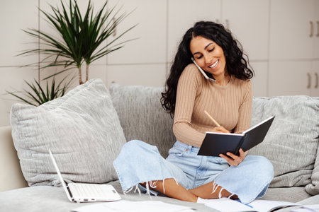 Busy woman talking by phone, write notes on notebook and working on laptop. Beautiful young arab woman plans meetings and organizes an event. Work as a manager remotely from home.の写真素材