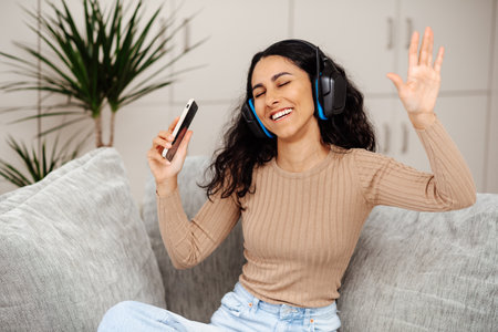 Portrait of a happy young Arab woman listening music with wireless headphones from music application on mobile smart phone in living room at condominium. Teen dancing listening to favorite playlistの写真素材