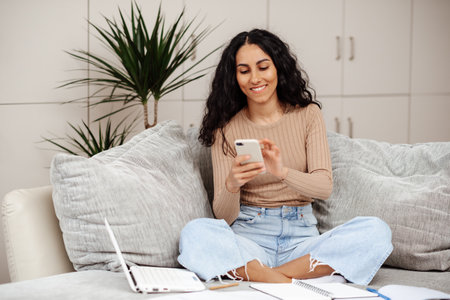 Smiling millennial mixed race woman looks at phone, reads message, has meeting, work with laptop in living room interior. App for communication remotely, study, freelance, good news at homeの写真素材