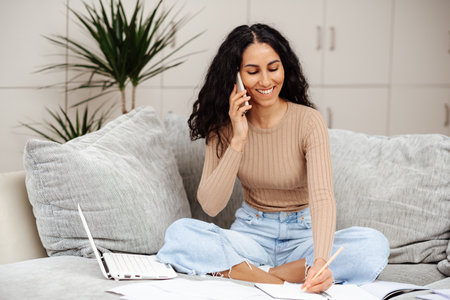 Education concept. Happy middle eastern woman making call about task, studying at home with laptop, taking notes while watching webinar or lesson. Writes homework in a notebook sitting on the sofaの写真素材