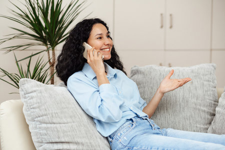 A beautiful young woman of Arab appearance sits on a sofa in a cozy home environment and talks on the phone. The girl is having a pleasant conversation, smiling brightly and gesturの写真素材