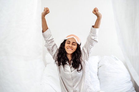 Girl sitting on bed after waking up in morning, stretching arms up and smilingの写真素材