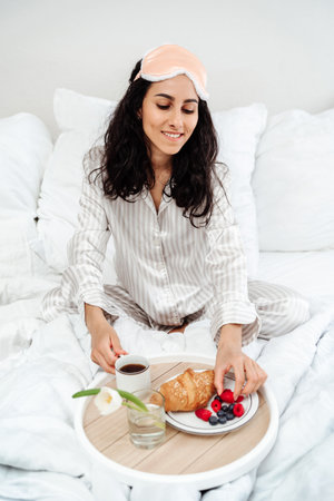 Cheerful tender female in white sleepwear sitting with cup of coffee on bed and having breakfast while enjoying morning in bedroom at home. Woman of Arab appearance and takes a cup of coffee and berryの写真素材