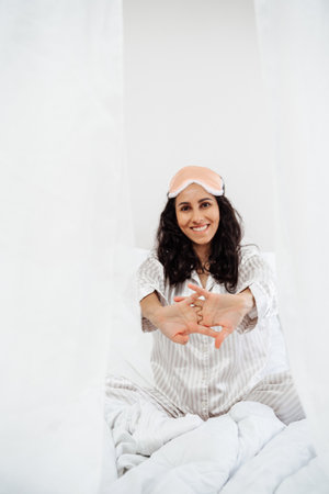 A woman of Eastern appearance woke up in a snow-white bed. The girl pulls her hands forward with her fingers crossed smiling brightly. Morning pull-ups are fun and the best start to the day.の写真素材