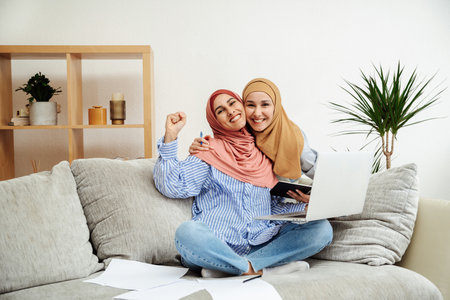 Two Arab women in hijabs successfully completed the exams. The girls smile broadly and celebrate the victory. Successful completion of distance learning. Training is over and they admiringly say yesの写真素材