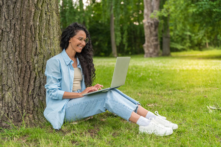 Happy Middle Eastern female student using laptop computer outdoors in the park. Girl learning tasks for preparing for exams. Woman with curly dark hair studying material for universityの写真素材