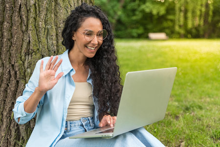 Pretty young of Hispanic woman sitting at park and having a video conference call on her laptop. Female sitting on grassy lawn having a video call on laptop. Girl in glasses with long curly hairの写真素材