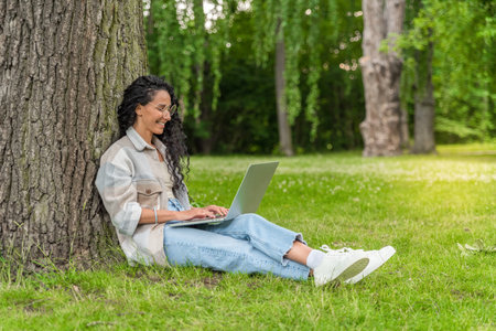 E-learning Concept. Arab Female Student With Laptop Sitting Under Tree In Park, Smiling Young Middle Eastern Woman Study Online On Computer While Relaxing On Green Lawn Outdoors, Copy Spaceの写真素材