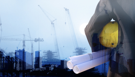 person construction worker man  or engineer holding in hands blueprint and yellow helmet for workers security on background of new highrise apartment buildings and construction cranes on backgroundの写真素材