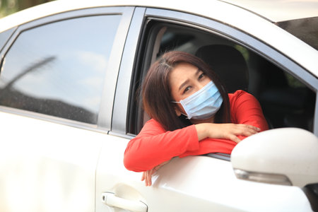 Young woman with medical protective mask on her face is driving a car, she looks into camera and View from outside , Protect yourself.の写真素材