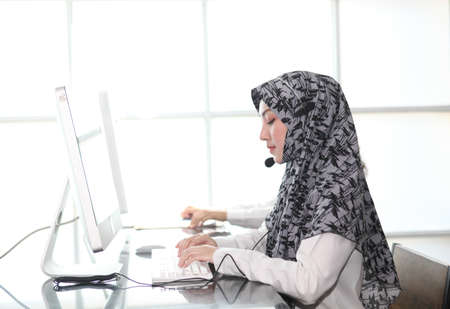 Asian Muslim female  with headsets working in a call centre,Call center agent with headset working on support hotline in modern office. Call center worker accompanied by her team. speaking to partnerの写真素材