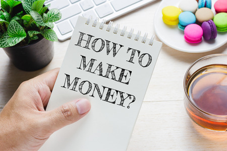 Man holding HOW TO MAKE MONEY message on book and keyboard with a hot cup of tea, macaroon on the table. Can be attributed to your ad.の写真素材