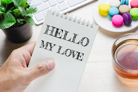 Man holding HELLO MY LOVE message on book and keyboard with a hot cup of tea, macaroon on the table. Can be attributed to your ad.の写真素材
