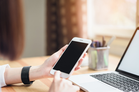 woman holding smartphone and using laptop on table in office room on windows with trees and nature background, for graphics display montage. Take your screen to put on advertising.の写真素材