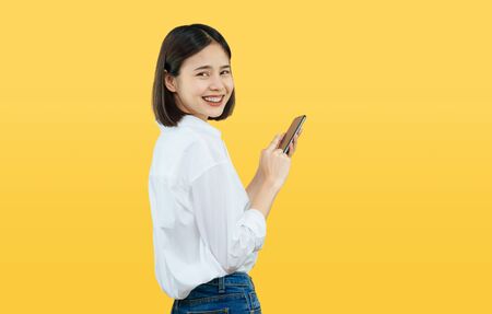Happy smiling Asian woman with holding smart phone on yellow background. copy space for put advertisement.の写真素材