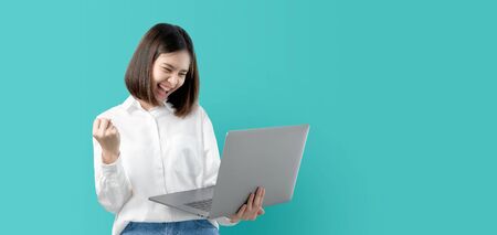 Young Asian woman smiling holding laptop computer with fist hand and excited for success on light blue background.