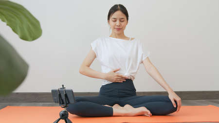 Asian women wearing sportswear and training online course yoga on smartphone in living room at home. wellness concept.の写真素材