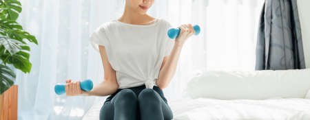 Asian woman sitting on the bed and doing exercises with small blue dumbbells, Warm-up the body in the morning. wellness concept.の写真素材