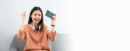 Excited young Asian woman wear white headphone on the head and playing games on smartphone in the living room at home.の写真素材